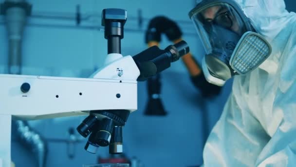 Ένας άντρας με στολή επικίνδυνων υλικών χρησιμοποιεί μικροσκόπιο για ανάλυση. — Αρχείο Βίντεο