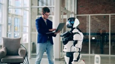 İş adamı dizüstü bilgisayar tutuyor ve modern bir robotla konuşuyor.