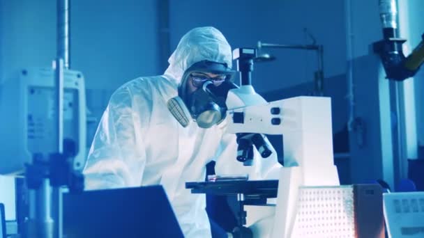 Εργαστηριακός εργάτης με στολή πιτσιλίσματος χειρίζεται μικροσκόπιο. — Αρχείο Βίντεο