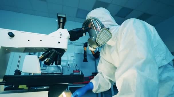 Ειδικός σε μια στολή πιτσιλίσματος εργάζεται με ένα μικροσκόπιο στο εργαστήριο — Αρχείο Βίντεο
