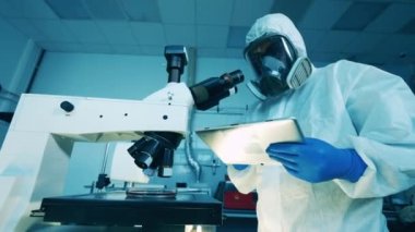 Tehlikeli madde giysisi giymiş bir bilim adamı laboratuvarda mikroskop kullanıyor.