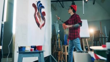 Resim stüdyosunda dövmeli bir adam resmin üzerine boya serpiyor.