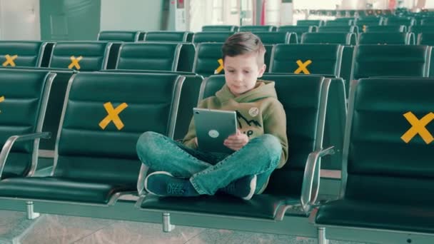 Мальчик сидит в пустом зале вылета и пользуется планшетом — стоковое видео