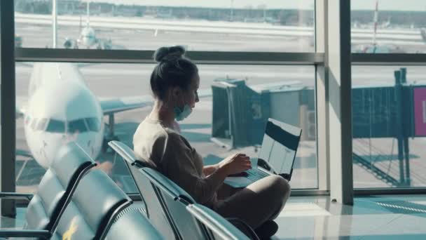 Женщина надевает маску для лица во время использования ноутбука в аэропорту — стоковое видео