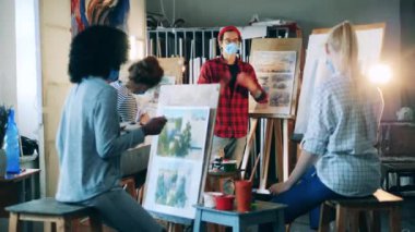 Yüz maskeli bir resim öğretmeni öğrencilerine resimleri anlatıyor.