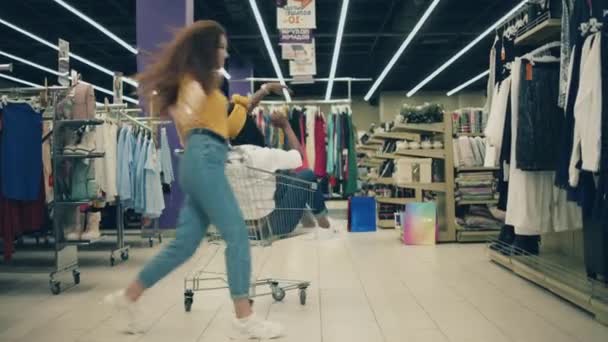 Compras felizes e divertidas. Duas senhoras bonitas estão dançando com um carrinho em uma loja de roupas — Vídeo de Stock