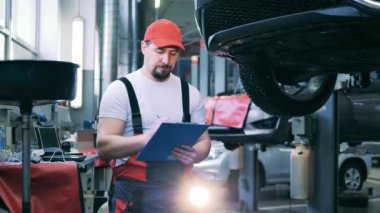 Araba tamircisi bir aracı aşağıdan inceliyor ve notlar alıyor.