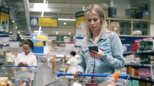 Una mujer está navegando por su teléfono mientras hace compras en el supermercado. Mujer en un supermercado, concepto de consumismo. — Vídeos de Stock