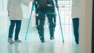 Doktorlar dış giysili bir adama koltuk değnekleriyle yürümesi için yardım ediyorlar.