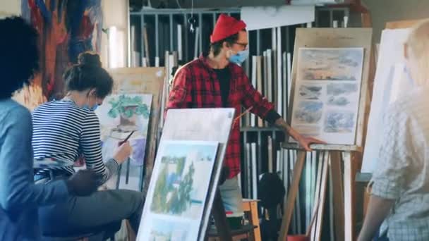 Pintor masculino em uma máscara facial está explicando pintura para estudantes do sexo feminino — Vídeo de Stock
