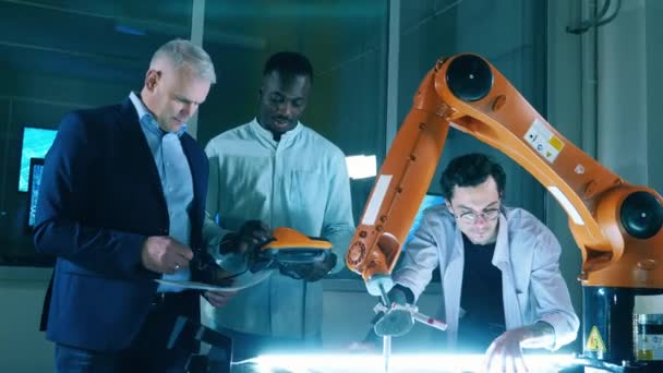 Οι άνδρες ερευνητές μιλούν ενώ λειτουργούν ένα ρομποτικό χέρι. Ομάδα μηχανικών που εργάζονται στο σύγχρονο καινοτόμο γραφείο. — Αρχείο Βίντεο