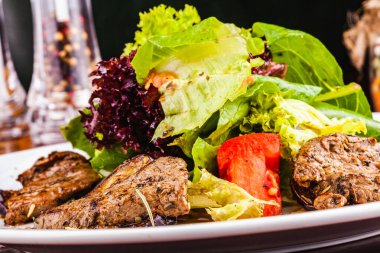 Beyaz tabakta patlıcanlı salata, biftek, marul ve domates.