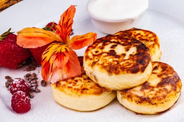 Kahvaltıda. Süzme peynirli krep, şurup taze böğürtlenli krep ve çilekli çilek, beyaz tabakta ekşi krema. Rus ya da Ukrayna mutfağı.