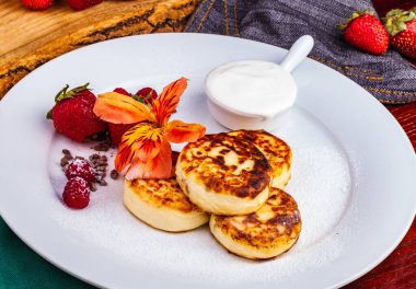 Kahvaltıda. Süzme peynirli krep, şurup taze böğürtlenli krep ve çilekli çilek, beyaz tabakta ekşi krema. Rus ya da Ukrayna mutfağı.