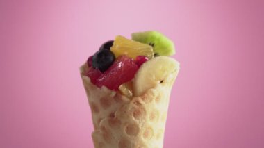 El, maşayla meyve salatasına yaban mersini koyar. Waffle külahında yabanmersinli taze ve sulu meyve karışımı pembe arka planda döner. Vejetaryen gıda konsepti.