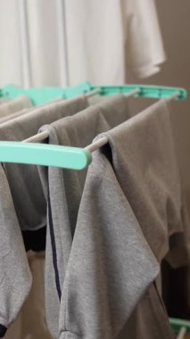 Giysiler parlak aydınlatılmış bir odanın içindeki kurutma askısına asılır. Yumuşak gri elbiseler yavaşça sallanıyor, basit bir çamaşır günü rutinini sergiliyorlar..