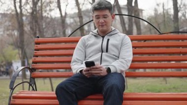 Smiling asian man using mobile phone while sitting on bench in public place. Portrait of cheerful person in glasses and light hoodie wearing modern smartwatch texting message on his smartphone.