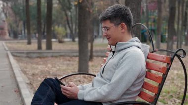 Gülümseyen orta yaşlı bir adam sonbahar parkında bankta oturup akıllı telefonuna bakıyor. Modern dijital yaşamı, iletişimi, teknoloji kullanımını ve boş zamanı yansıtan doğal belgesel tarzı bir an.