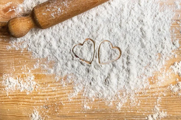 Yılın en güzel günü için kurabiyeleri hazırla: sevgililer günü. Tahta masaya saçılmış un üzerine basılmış iki kalp. Sevgiyi yay!