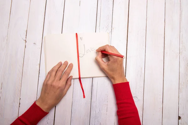 Female hands writing a lovely message on her notebook. The touch of red from the sweater and the pencil recall the Valentine color. White wooden background with empty space for text.