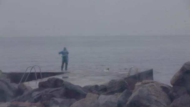 De eenzame visser op een stenen pier in regenachtig weer vangt vis — Stockvideo