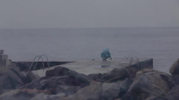 Одинокий рыбак на каменном пирсе в дождливую погоду ловит рыбу — стоковое видео