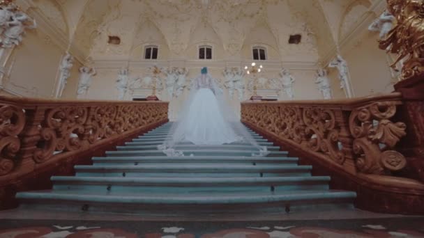 La novia en el vestido lujoso blanco con el nudo ancho grande se levanta sobre la escalera de la gran sala dorada — Vídeos de Stock