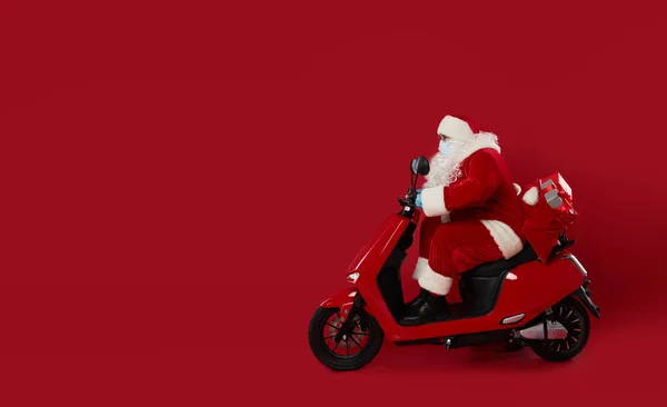 Elderly Santa in a protective mask and gloves on a retro motorcycle delivering a bag of gifts during the coronavirus quarantine. Red studio background.