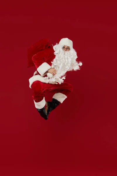スーパーヒーローポーズの喜ばしいサンタクロースは 赤い背景の贈り物で高くジャンプします クリスマスと新年の休日と販売のための概念 — ストック写真