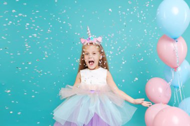 Tek boynuzlu at kızı. Zarif tül elbise ve tek boynuzlu at saç bandı takan neşeli kız doğum günü partisini kutluyor ve arka planda renkli konfeti üflüyor.
