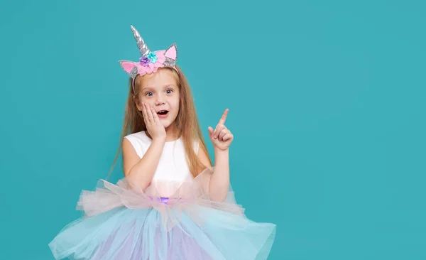 Unicorn girl. Wow face. Cute child girl in elegant tulle dress and unicorn headband celebrates birthday  party on colored background. Points to the right