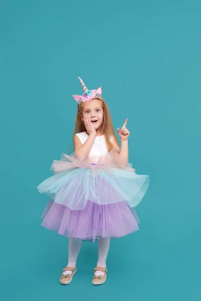 Unicorn girl. Funny face. Cute child girl in elegant tulle dress and unicorn headband celebrates birthday  party on colored background. Points to the right