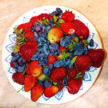 Bir tabak dolusu olgun kırmızı çilek ve koyu mor hanımeli böğürtlen, yeni toplanmış.