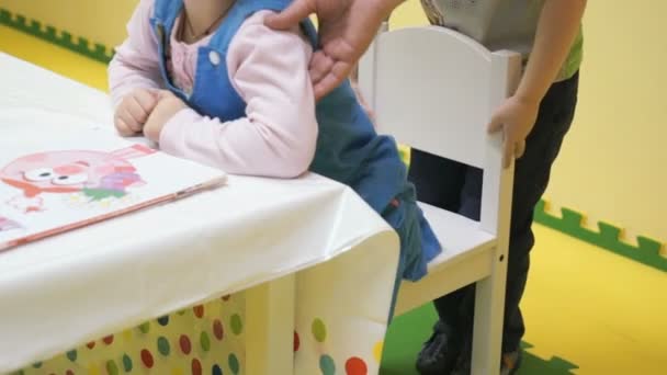 Una niña pequeña ayuda a un niño a sentarse en una silla — Vídeo de stock