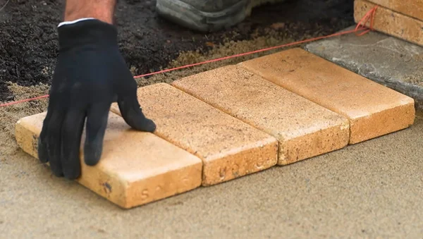 Bir işçi, bronzlaşmış tuğlaları düz bir çizgiye, pürüzsüz ve yeni hazırlanmış bir yüzeye dikkatlice yerleştirir. Eldivenler korunmak için takılıyor ve aletler yakınlarda, devam eden inşaat faaliyetlerini gösteriyor..