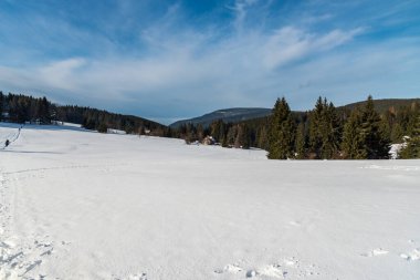 Kış manzarası karla kaplı çayır, az sayıda yürüyüşçüyle yürüyüş yolu, arka planda birkaç izole ev ve tepeler ve Çek Cumhuriyeti 'nin Moravskoslezske Beskydy dağlarındaki Lysa hora tepesi