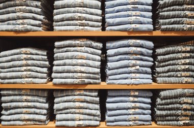 Perakende mağazasındaki tahta rafta düzgünce katlanmış çizgili gömlekler. Her giysi, verimli bir envanter yönetimi ve küratörlü moda koleksiyonunu temsil eden görünür bir boyut etiketi (S, M, L) içerir..