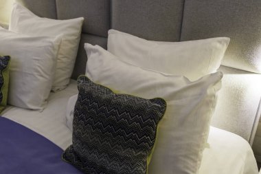 Koyu renkli, desenli, yeşil kenarlı bir yastığın açılı ve modern yatak odasındaki gri kumaştan bir yatak başlığına yaslanmış beyaz yastıklar..
