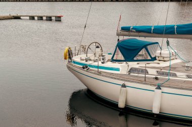 Sakin bir marinaya yanaşmış beyaz ve mavi bir yelkenlinin pruvası, arka planda ahşap bir güverte, karanlık su ve yüzen jettiler var..
