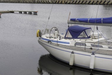 Mavi aksanlı beyaz bir yelkenlinin ön kısmının yakın çekimi. Direksiyon simidi ve ahşap güvertesi olan sakin bir limana yanaşmış..