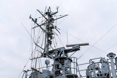 Karmaşık, metalik direğin düşük açılı yakın çekimi ve bulutlu bir gökyüzüne karşı büyük gri bir askeri geminin antenleri..