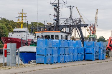 Kenetlenmiş bir balıkçı teknesi ya da servis teknesi, ön planda istiflenmiş mavi plastik kaplar ve gri bir gökyüzünün altında sarı kargo vinçleri içeren yoğun bir sanayi limanı sahnesi..