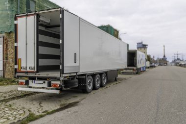 Bulutlu bir günde, endüstriyel bir liman bölgesinin asfalt yoluna park etmiş, kapısı açık, büyük beyaz soğutmalı yarı römorkun arka ve yan görüntüsü..