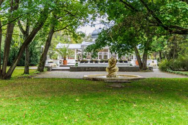 Çimenli ön planda dekoratif bir heykel çeşmesinin yer aldığı, bereketli ağaçlarla çerçevelenmiş camdan bir cephesi olan klasik bir beyaz park binası (pavyon / restoran)..