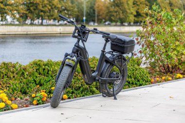 Arka kargo kutusu olan modern bir siyah yağ lastikli bisiklet (e-bisiklet), peyzajlı bir çalı sınırı ve bir şehir nehri veya kanalın yanındaki kaldırımlı bir yola park edilmiştir..