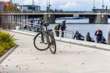 Temiz, asfalt bir setin üzerine park edilmiş bir bisiklet, bulanık bir arka plan ile çok sayıda insanın su kenarında balık tuttuğunu ya da toplandığını ve bir köprünün kanalı geçtiğini gösteriyor..