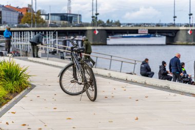 : Bir bisiklet asfalt bir şehrin rıhtımında sığ bir tarla derinliğinde duruyor. Birden fazla insan balık tutarken ve bir köprü uzaktaki suyu kaplarken motosikleti parlaklaştırıyor..