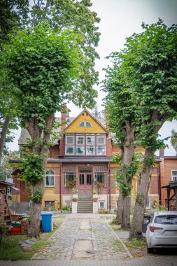 İki katlı eski bir ahşap bina, sarı bir cephe, geniş pencereler, ve uzun, budanmış ağaçlarla döşenmiş asfalt bir yürüyüş yolu..