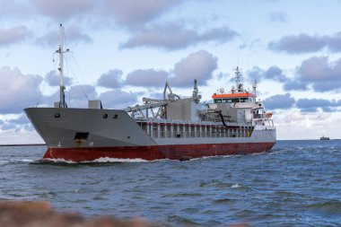 Orta büyüklükte bir kargo gemisi, dalgalı suda, geminin iskele tarafında ve dramatik, bulutlu bir gökyüzünün altında görünen dalgalı dalgalı sularla izleyicinin yanından geçiyor..