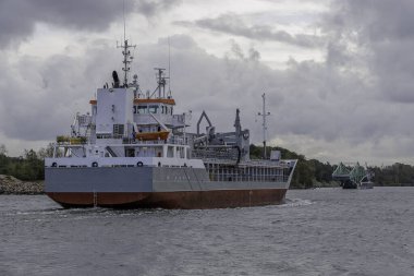 Orta büyüklükteki bir kargo gemisi kıç tarafı görünen bir kanaldan geçer, uzak, yeşil bir salıncak köprüsünü geçer ve ağır bulutlu bir gökyüzünün altında yol alır..